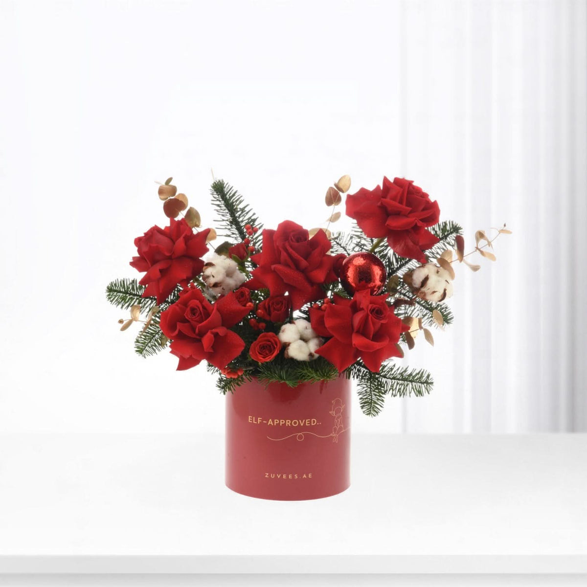 Vibrant red roses, pine, cotton, and ornament in festive red cylindrical box.