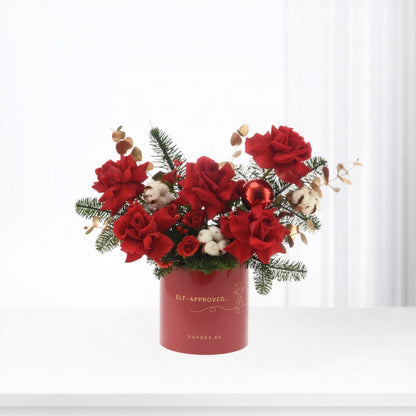 Vibrant red roses, pine, cotton, and ornament in festive red cylindrical box.