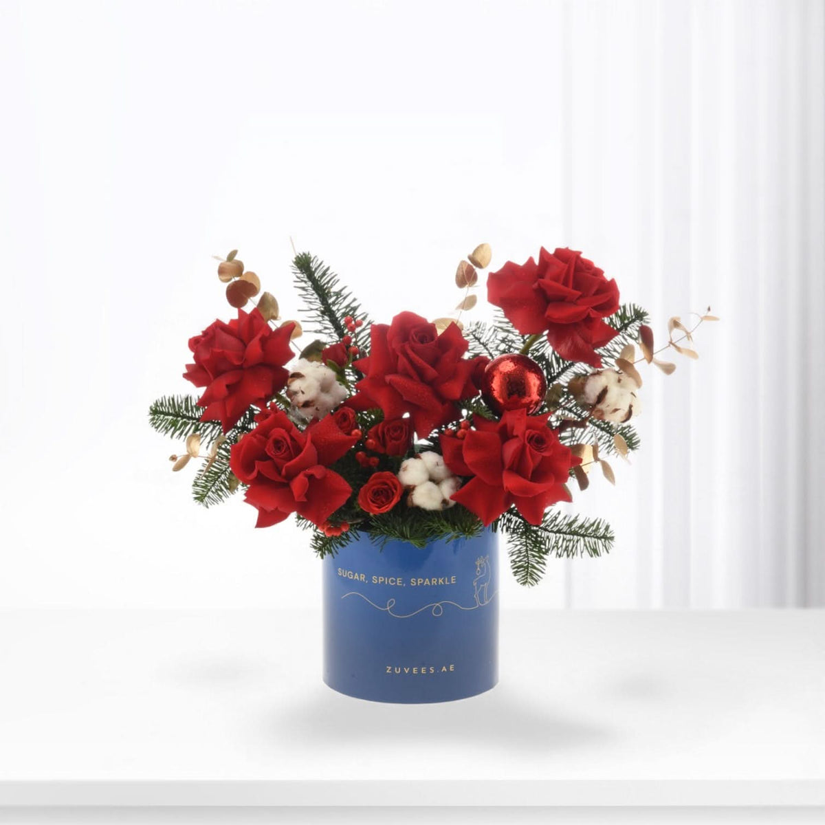 Vibrant red roses, cotton, pine, and a red ornament in a blue "Sugar, Spice, Sparkle" vase.