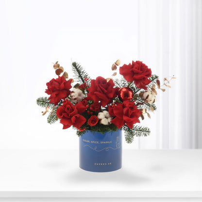 Vibrant red roses, cotton, pine, and a red ornament in a blue "Sugar, Spice, Sparkle" vase.