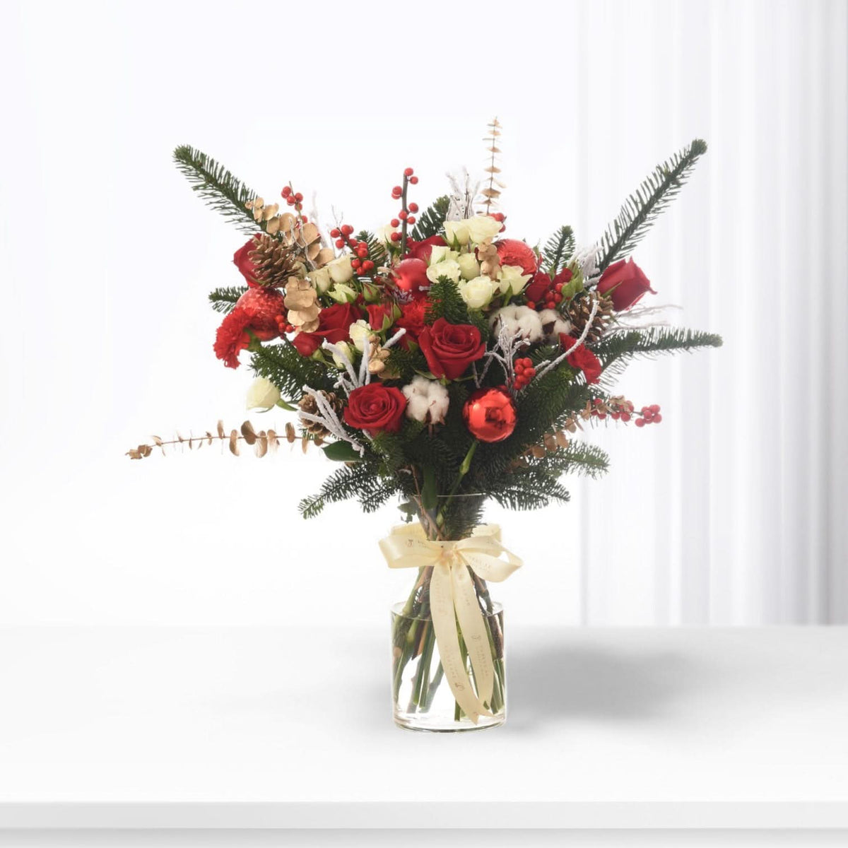 Vibrant red roses, berries, pine, and cotton in clear vase with cream bow.