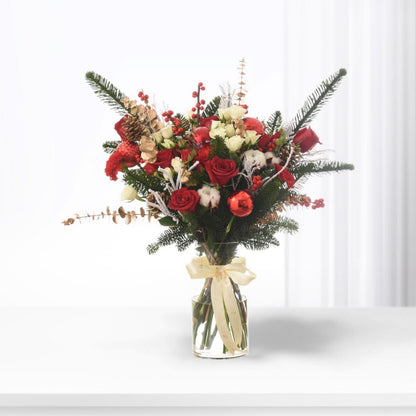 Vibrant red roses, berries, pine, and cotton in clear vase with cream bow.