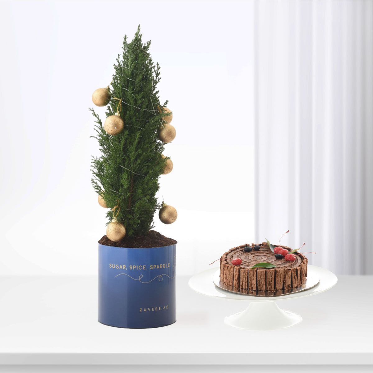 Blue pot Christmas tree with gold ornaments beside chocolate cake on white stand.