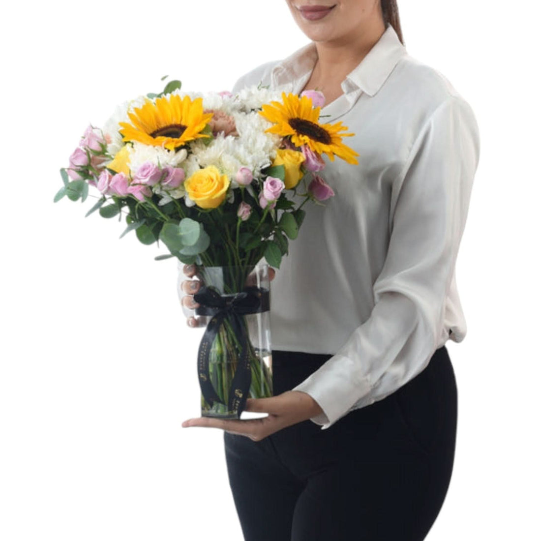 Woman holds vibrant sunflower, rose, and chrysanthemum bouquet in clear glass vase with black ribbon.