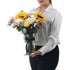 Woman holds vibrant sunflower, rose, and chrysanthemum bouquet in clear glass vase with black ribbon.