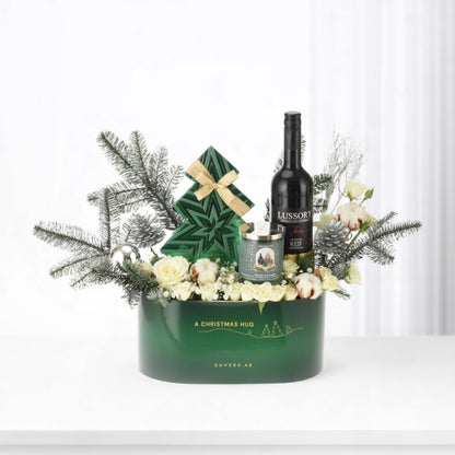 Green gift basket with white roses, pine, candle, and wine bottle.