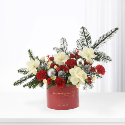 Red cylindrical vase holds festive white roses, red carnations, pine, and silver ornaments.