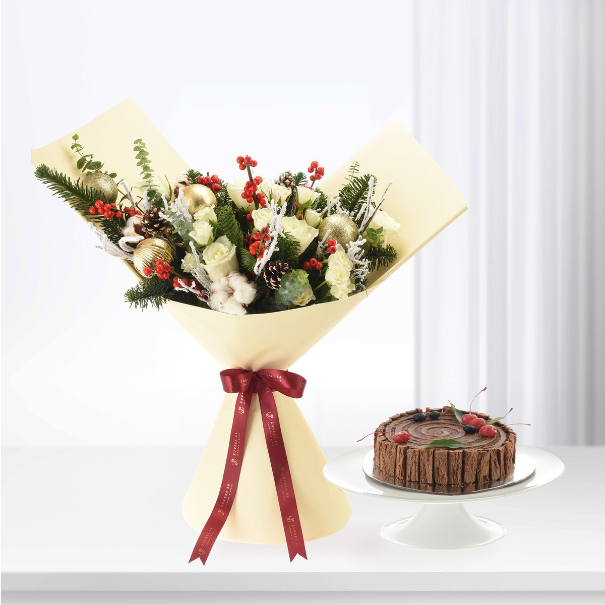 Festive white rose bouquet with gold ornaments, red berries, pinecones, beside chocolate log cake.