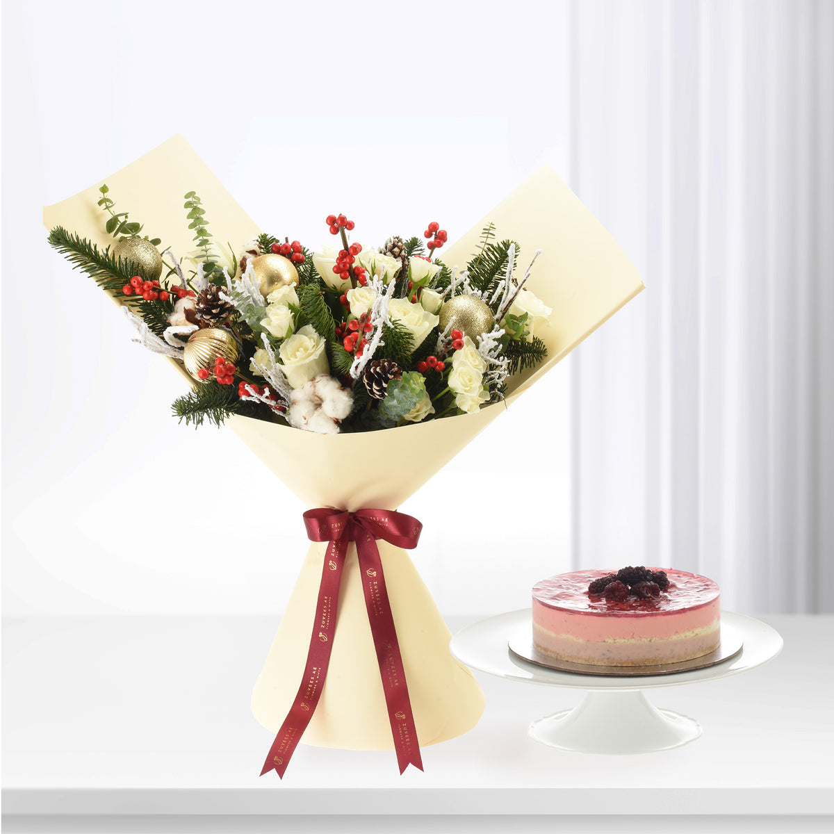 Elegant cream-wrapped floral bouquet with gold ornaments, red berries, and pine, beside a layered berry cake.