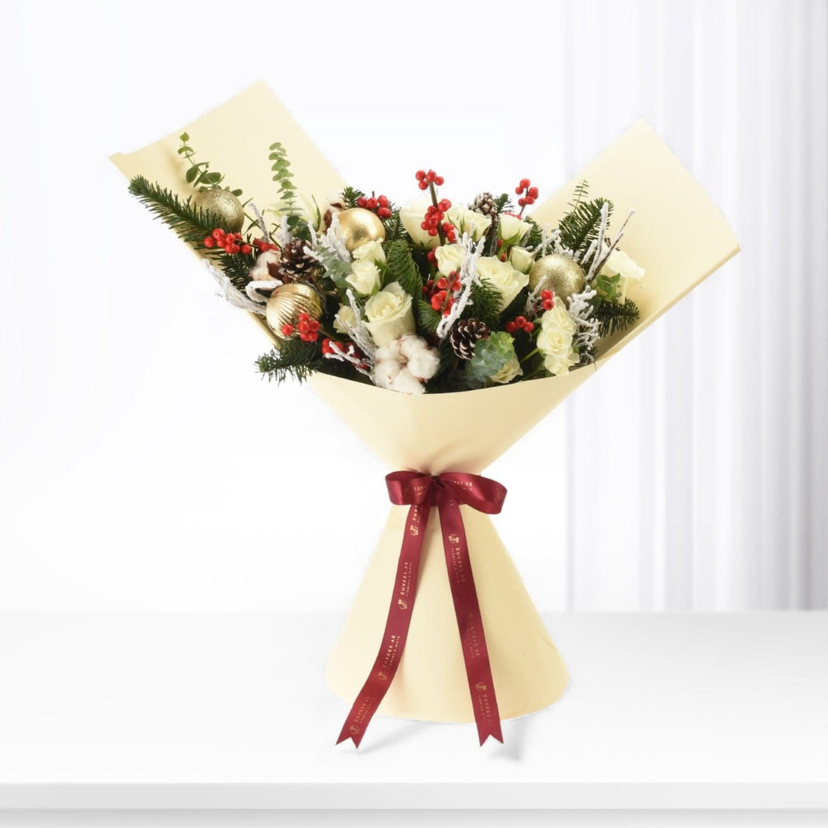 Cream-wrapped holiday bouquet with white roses, red berries, pine, gold ornaments, and burgundy ribbon.
