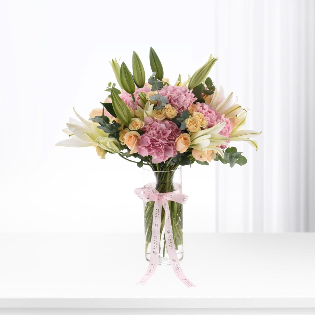 Pastel pink hydrangeas, white lilies, and peach roses in clear vase with pink ribbon.