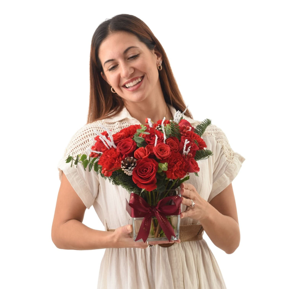 Woman smiles holding vibrant red roses and carnations in glass vase with burgundy bow.