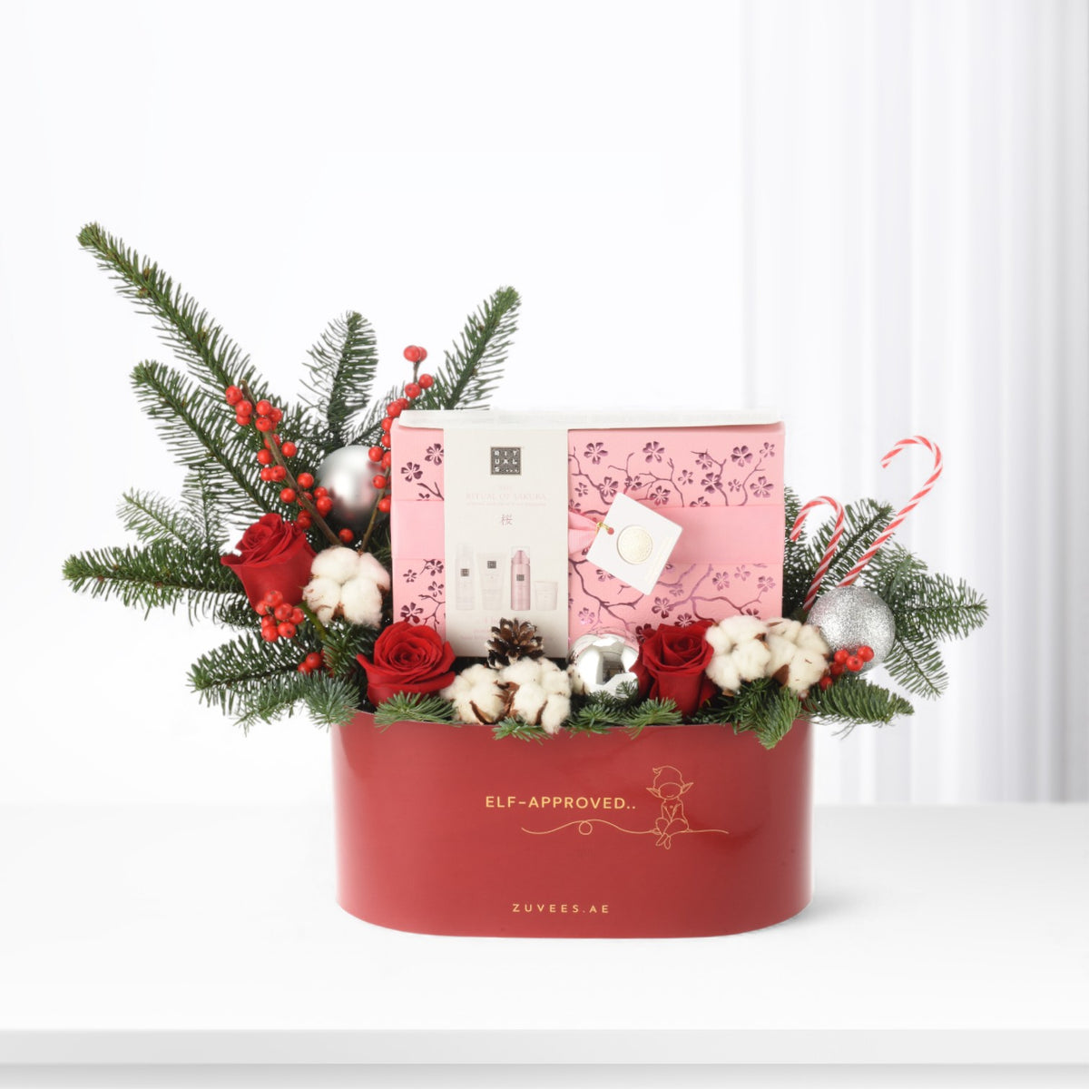 Festive red gift box with roses, pine, ornaments, and pink skincare set inside.