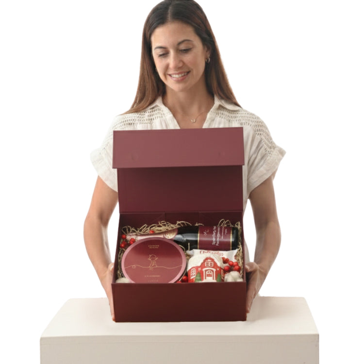 Woman smiles holding open burgundy gift box revealing wine, cookies, and festive treats.