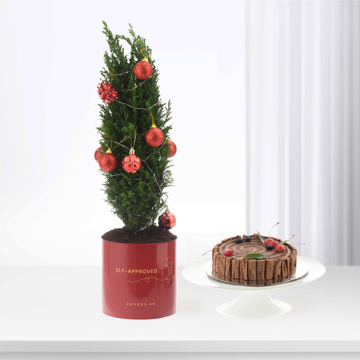 Mini Christmas tree in red pot with glitter ornaments beside chocolate yule log cake.