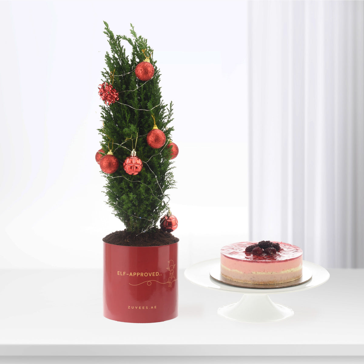 Red-ornamented mini tree in glossy pot beside layered berry cake.