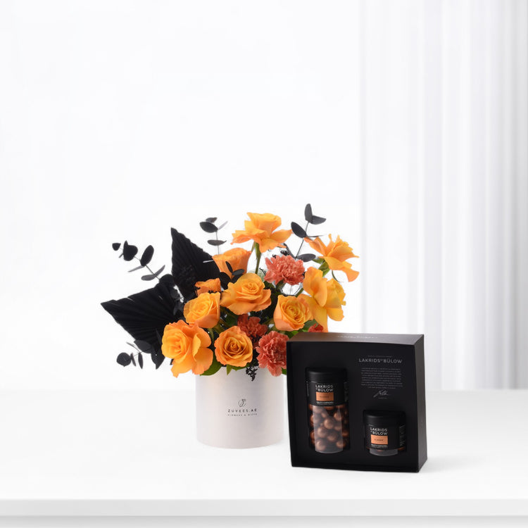 Vibrant orange roses and black foliage in white vase beside gourmet chocolate gift box.