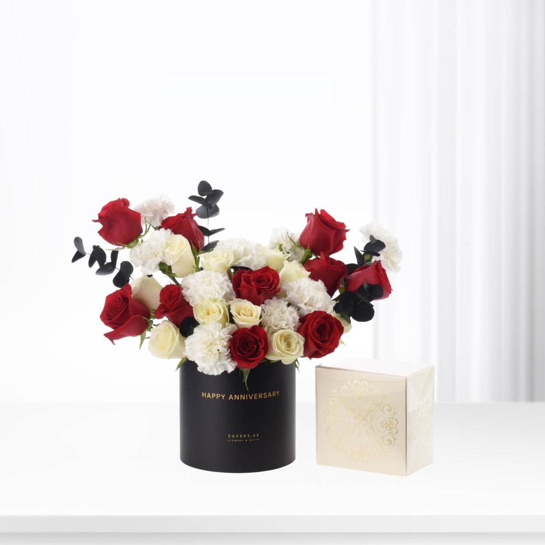 Elegant red, white roses and eucalyptus in black "Happy Anniversary" vase beside gold-embossed gift box.