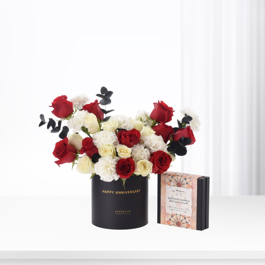 Elegant red and white roses with black eucalyptus in black "Happy Anniversary" vase, beside spice caramel gift box.