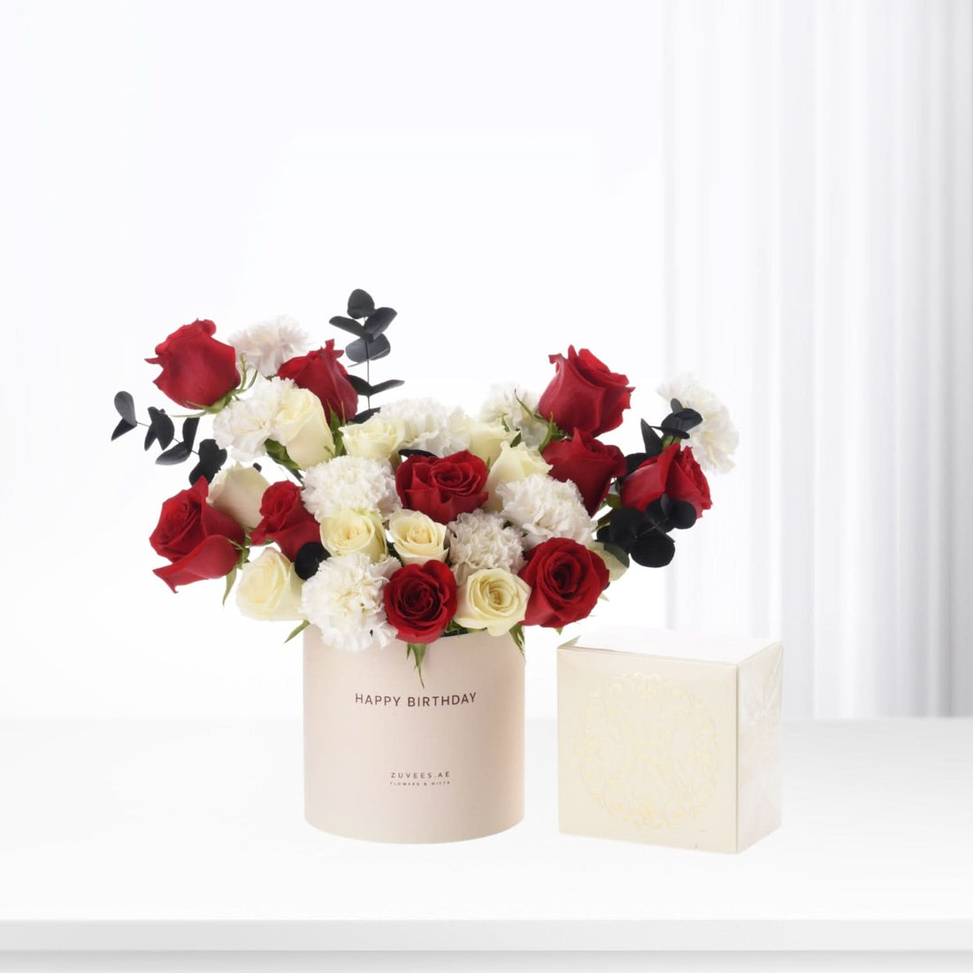 Elegant red, white roses and carnations in beige "Happy Birthday" cylinder beside ornate gift box.
