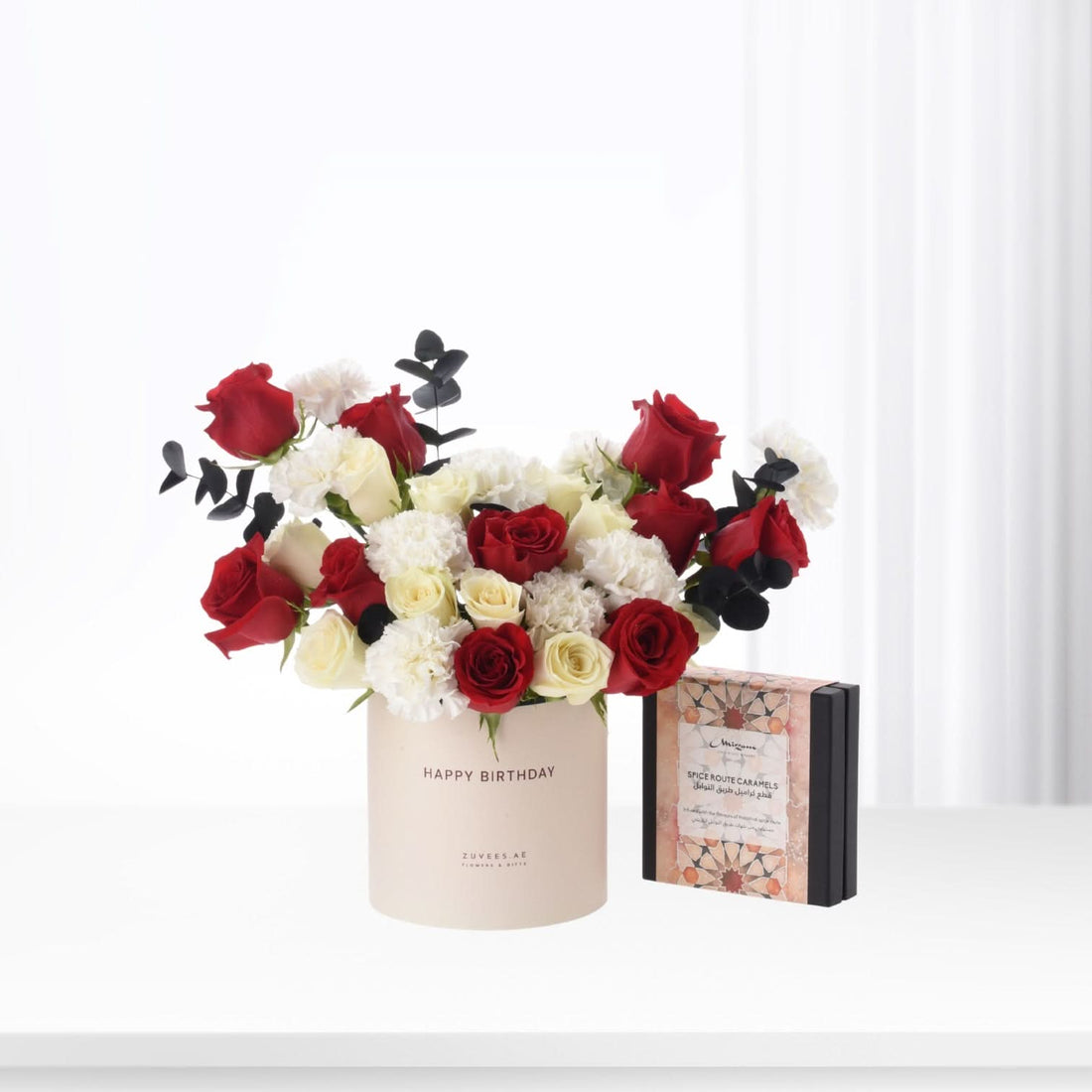 Red and white roses with black eucalyptus in beige "Happy Birthday" box beside caramel gift.