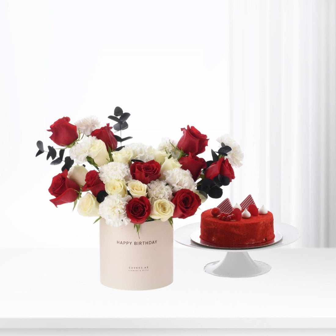 Red and white roses with black eucalyptus in beige box beside red velvet cake.