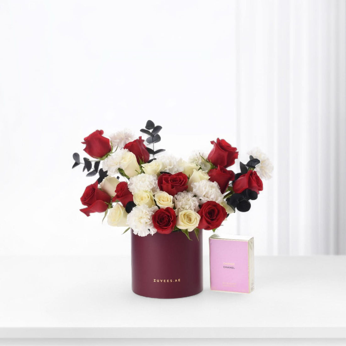 Luxurious red, white, and black rose bouquet in burgundy cylinder beside pink Chanel perfume.