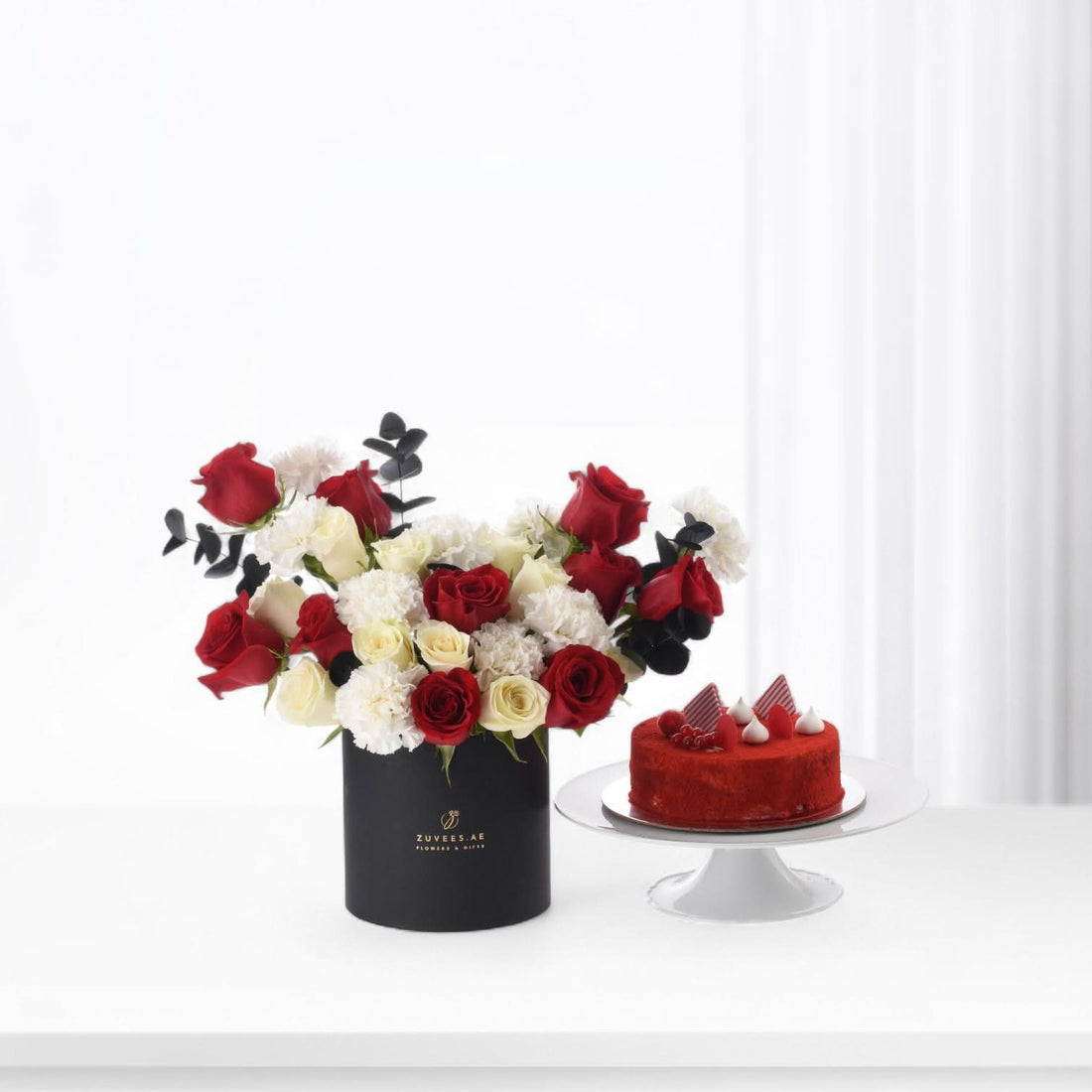 Elegant red and white roses with black eucalyptus, beside a red velvet cake on white stand.