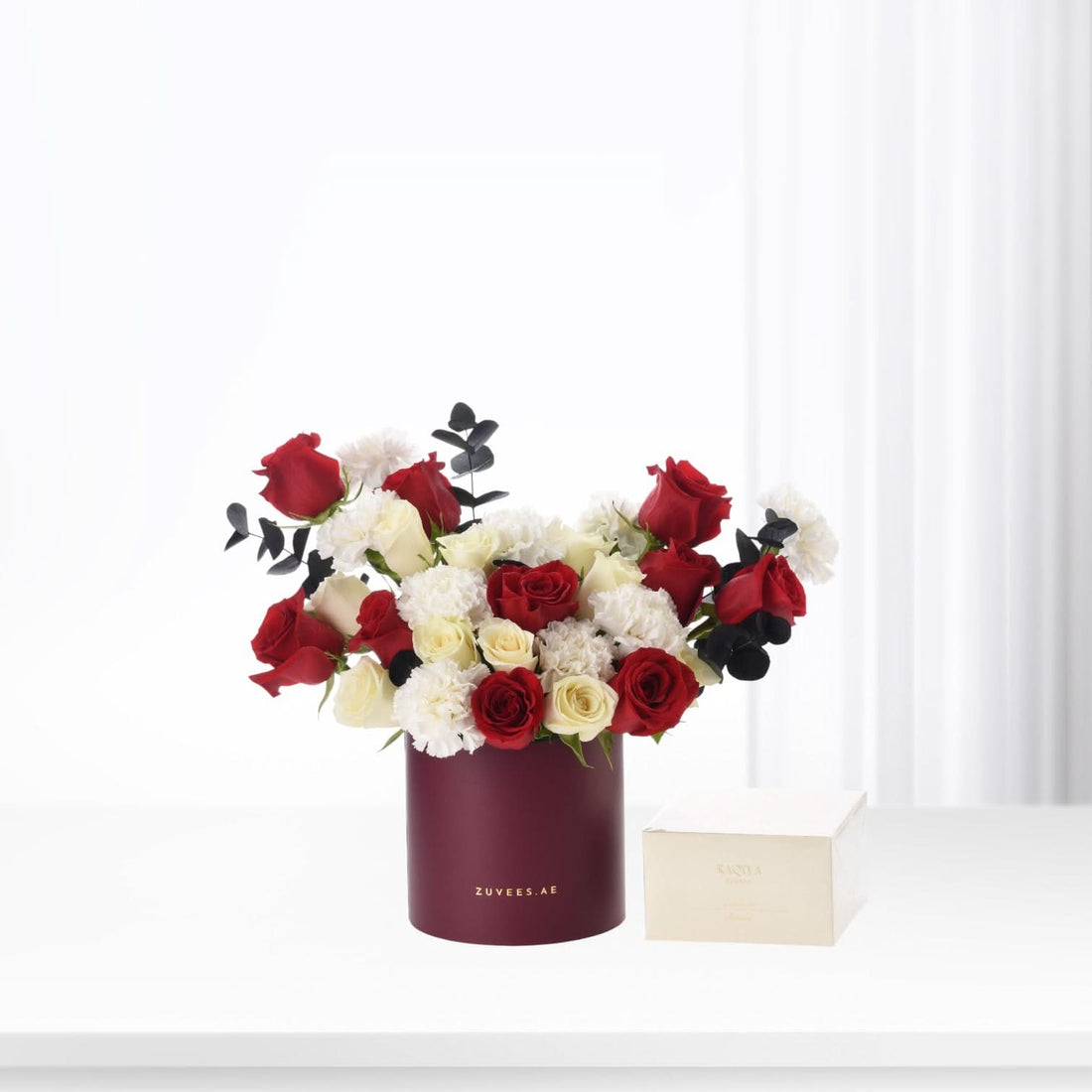 Vibrant red, white, and cream roses with black eucalyptus in a deep burgundy cylindrical vase.