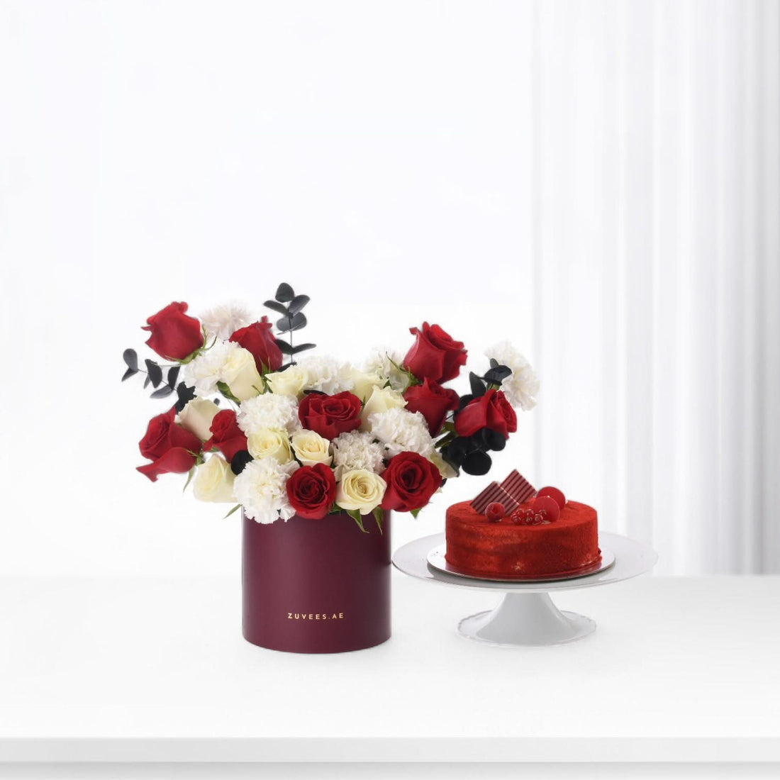 Red and white roses in burgundy vase beside red velvet cake on white stand.