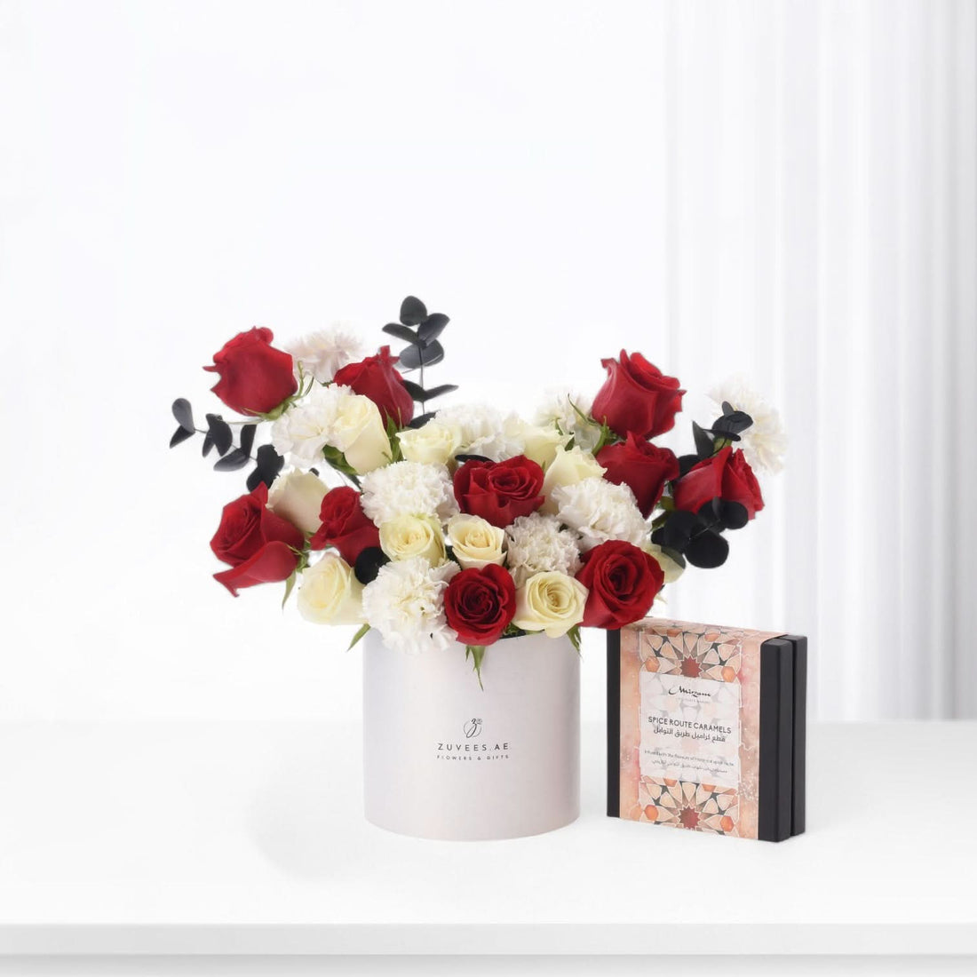 Elegant red, white roses and carnations in minimalist white box beside spice caramel gift.