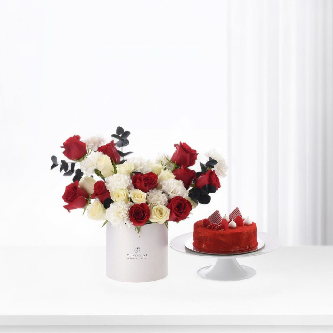 Red and white roses with carnations in white cylinder beside red velvet cake on pedestal.