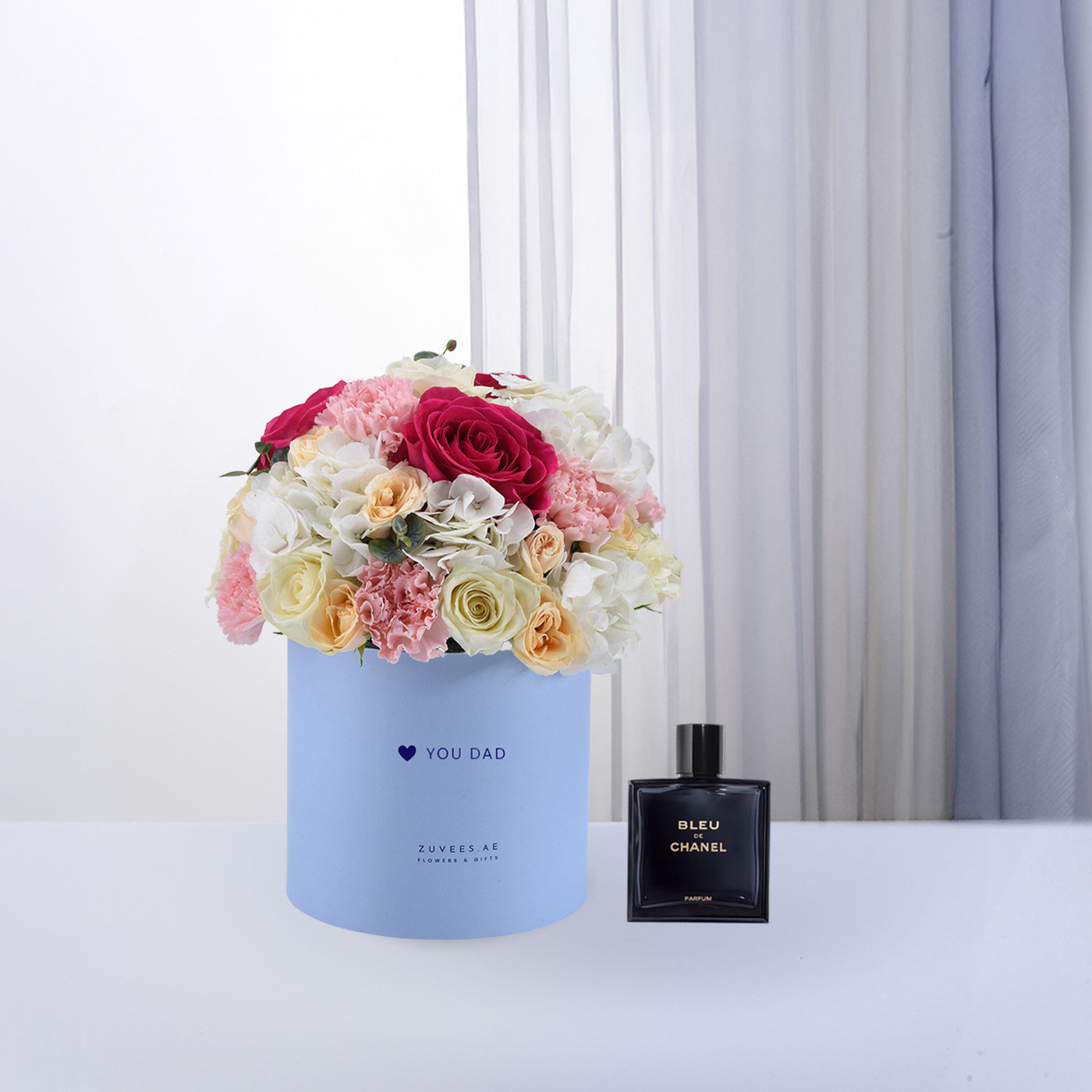 Pastel blue box with vibrant roses, carnations, and hydrangeas beside Chanel Bleu de Chanel perfume.