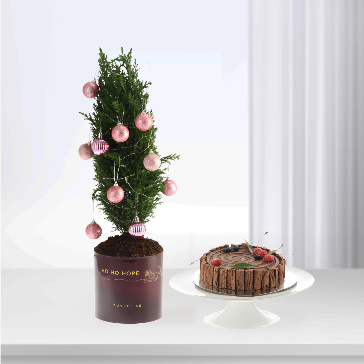 Pink baubles adorn a green tree in burgundy pot beside chocolate cake.