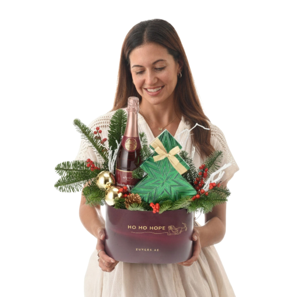 Woman smiles holding festive holiday gift basket with champagne, green tree, and red berries.