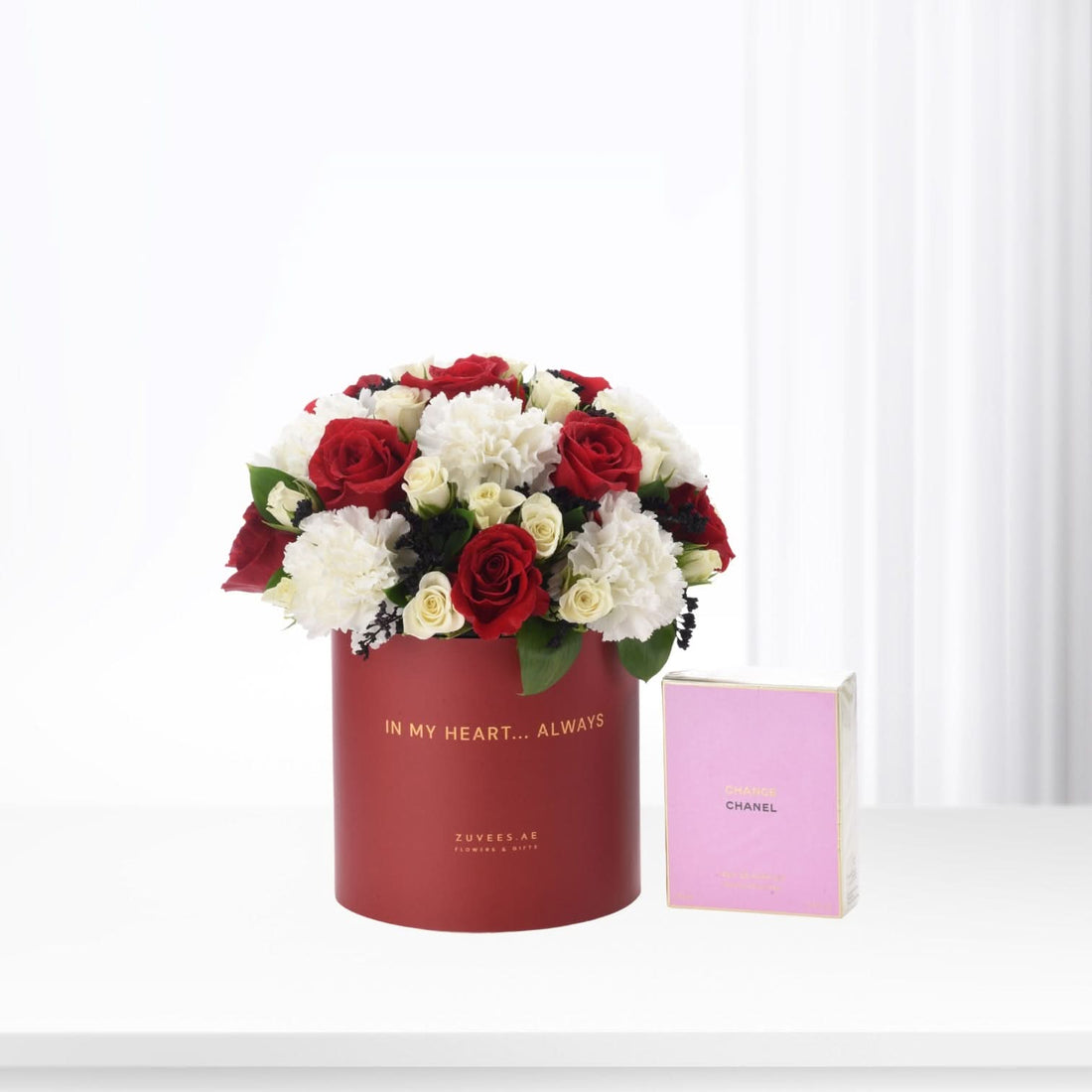 Red cylindrical box with "In My Heart... Always" text, holding red and white roses and carnations, beside pink Chanel Chance perfume box.