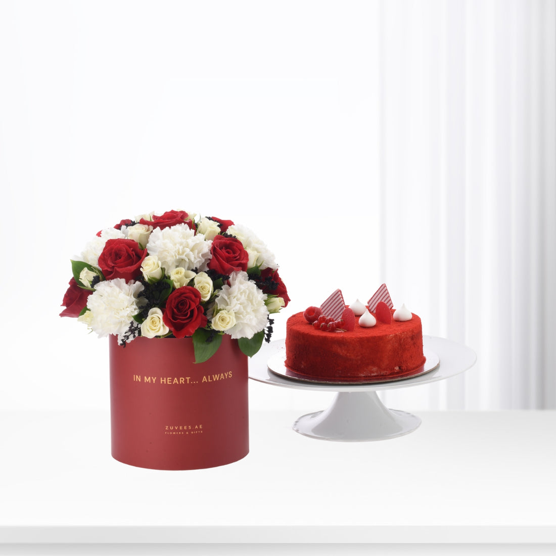 Red roses and white carnations in crimson box beside red velvet cake.