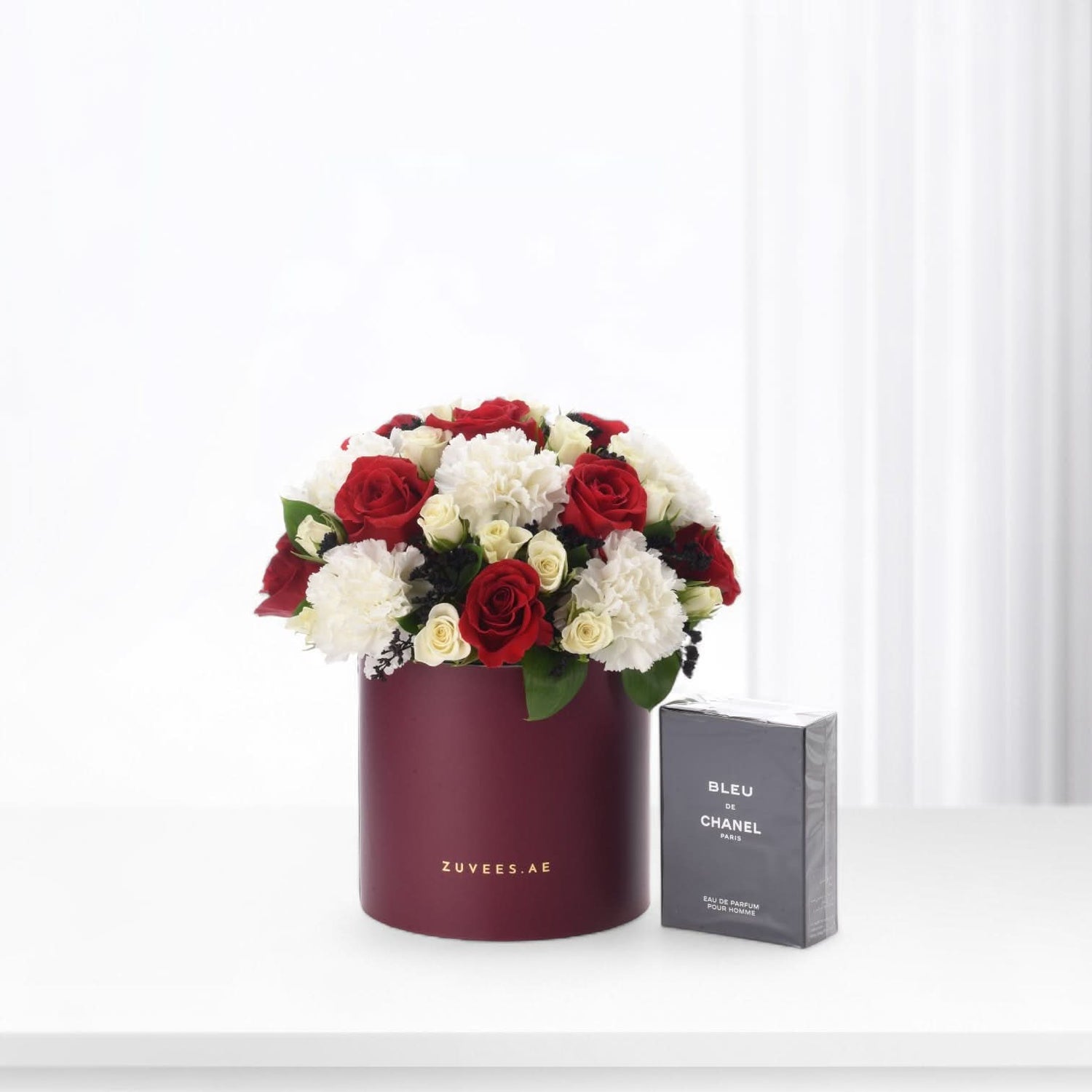 Elegant red and white roses in burgundy box beside Chanel Bleu perfume.