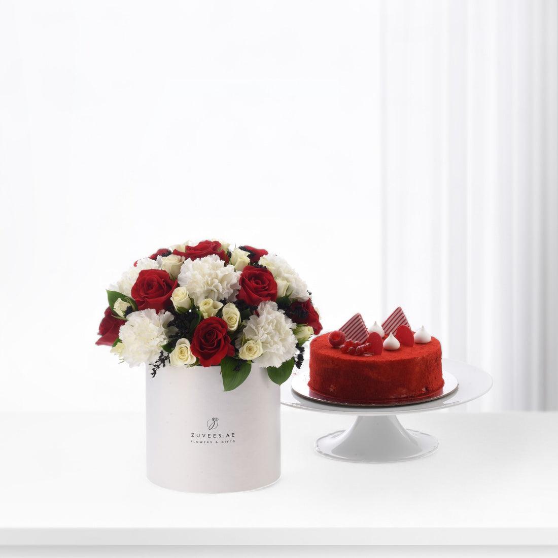 Red roses and white carnations in white box beside red velvet cake.