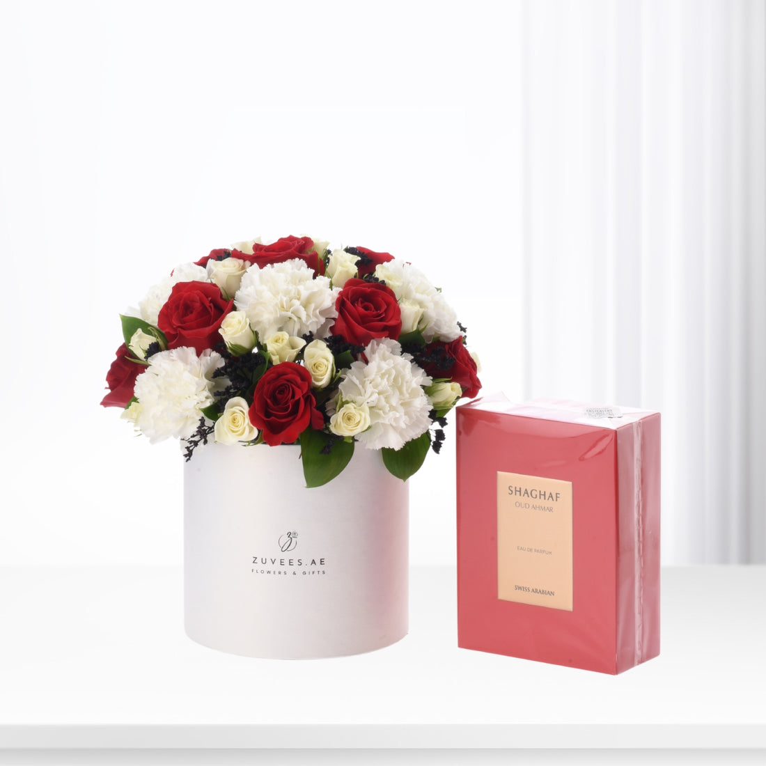Red and white roses with carnations in white box, beside pink perfume box.