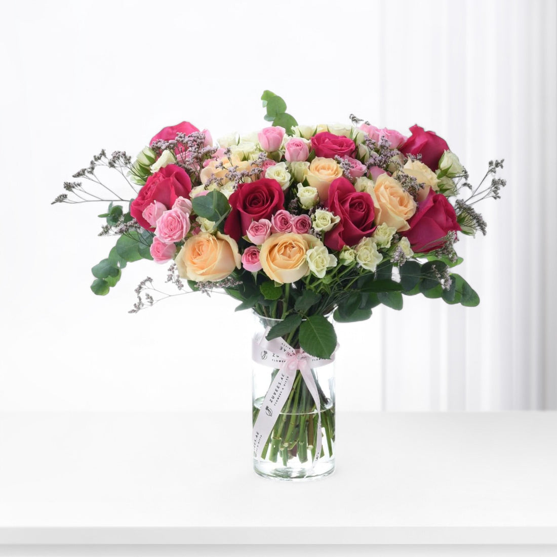 Vibrant multicolor roses in clear vase with pink ribbon, fresh greenery, and delicate filler blooms.