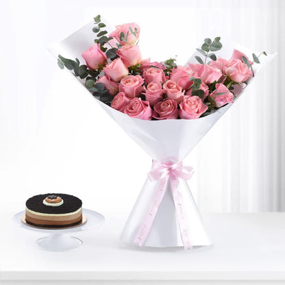 Pink roses bouquet with eucalyptus, white wrap, pink ribbon, beside chocolate cake.