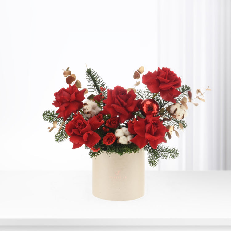 Vibrant red roses, cotton, pine, and a glitter ball in a cream vase.