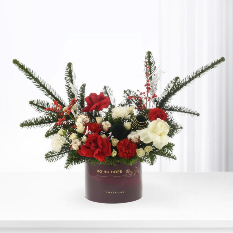 Festive red and white roses with pine, berries, gold accents in maroon "HO HO HOPE" vase.
