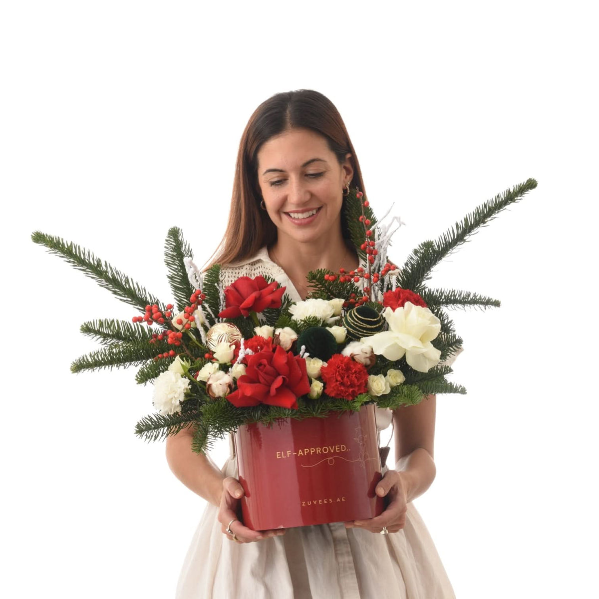 Woman holds festive red box with evergreens, red roses, white blooms, and berries.