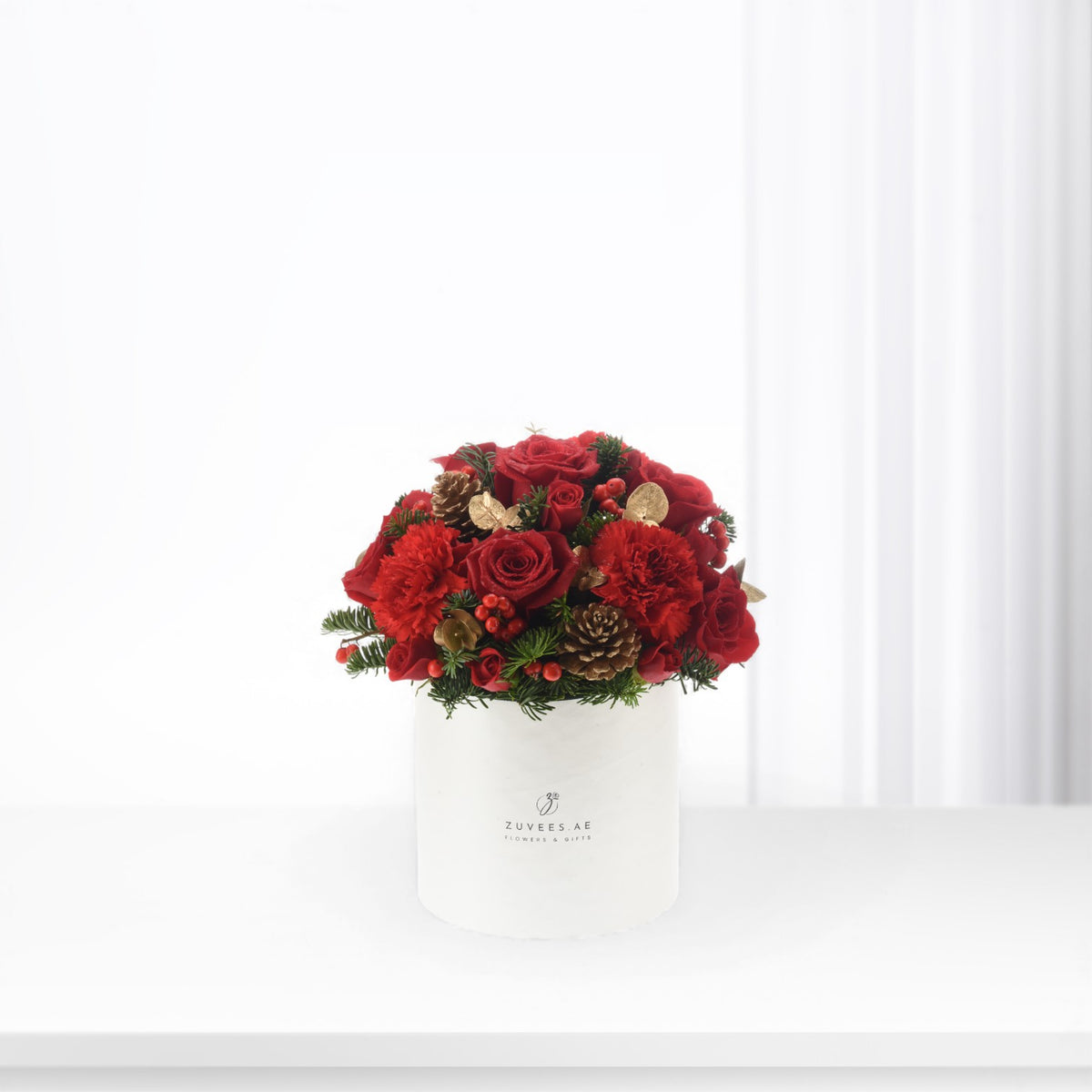 Vibrant red roses and carnations in white box with pinecones and gold accents.