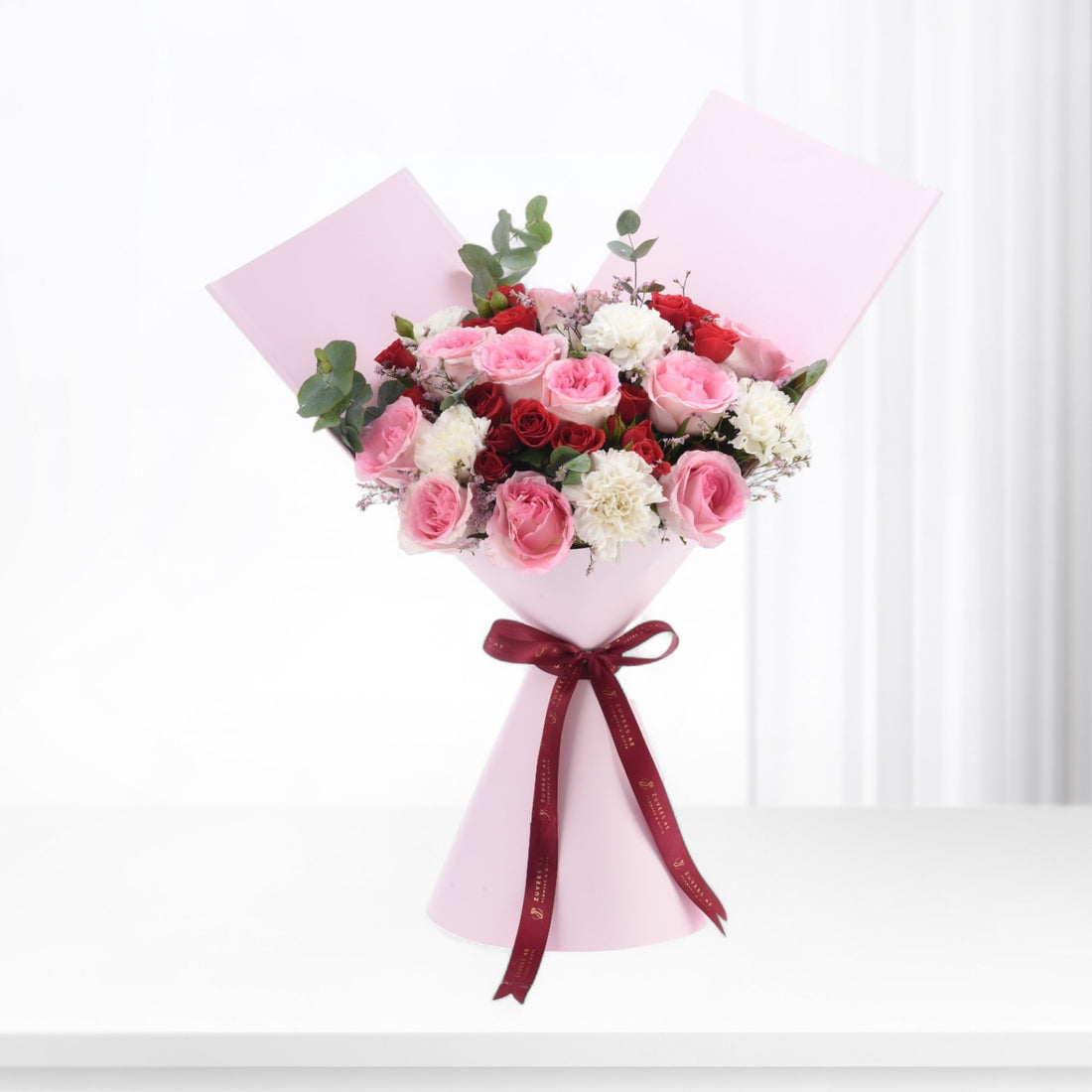 Pink-wrapped bouquet with red, pink, and white roses, accented by greenery and a burgundy ribbon.