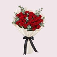 A luxurious hand-tied bouquet of vibrant red roses wrapped in cream silk paper, accented with lush green eucalyptus foliage. The arrangement is finished with a black ribbon bow, against a soft neutral background.