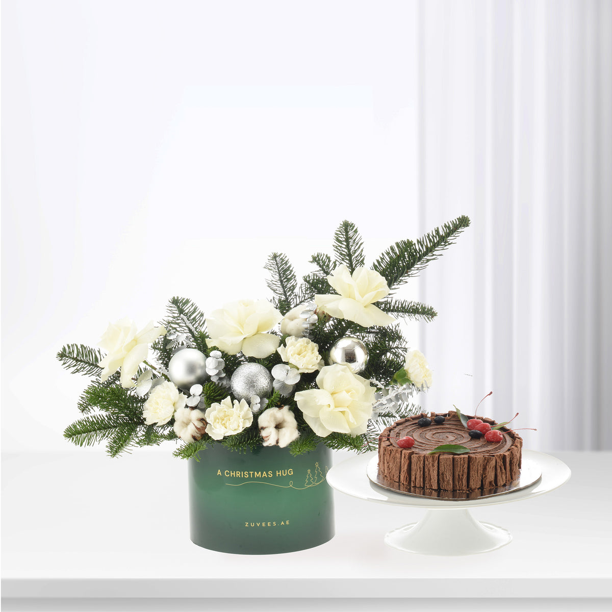 Elegant green holiday bouquet with white blooms and silver ornaments beside chocolate cake.