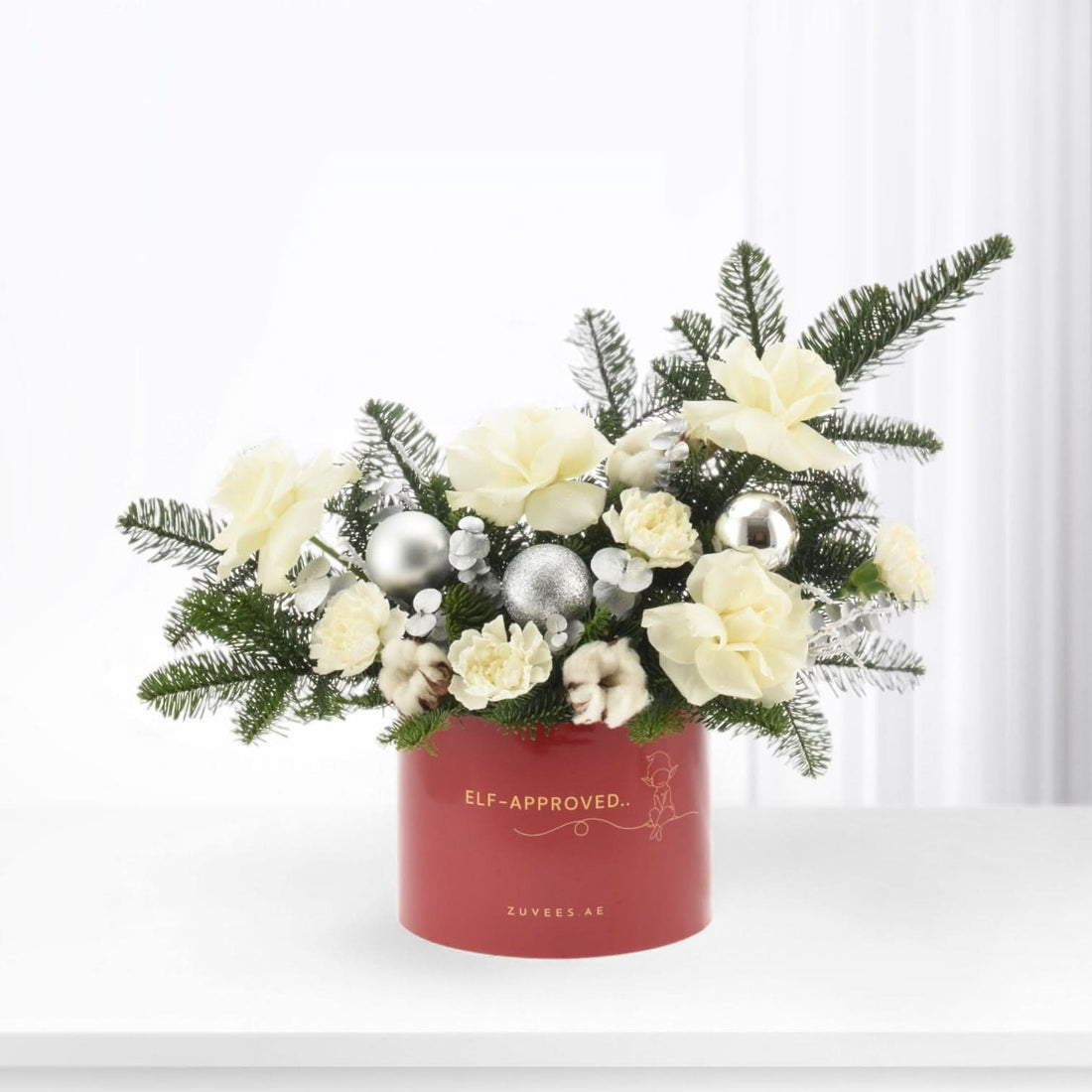 Red holiday floral arrangement with white blooms, silver ornaments, and pine in festive container.